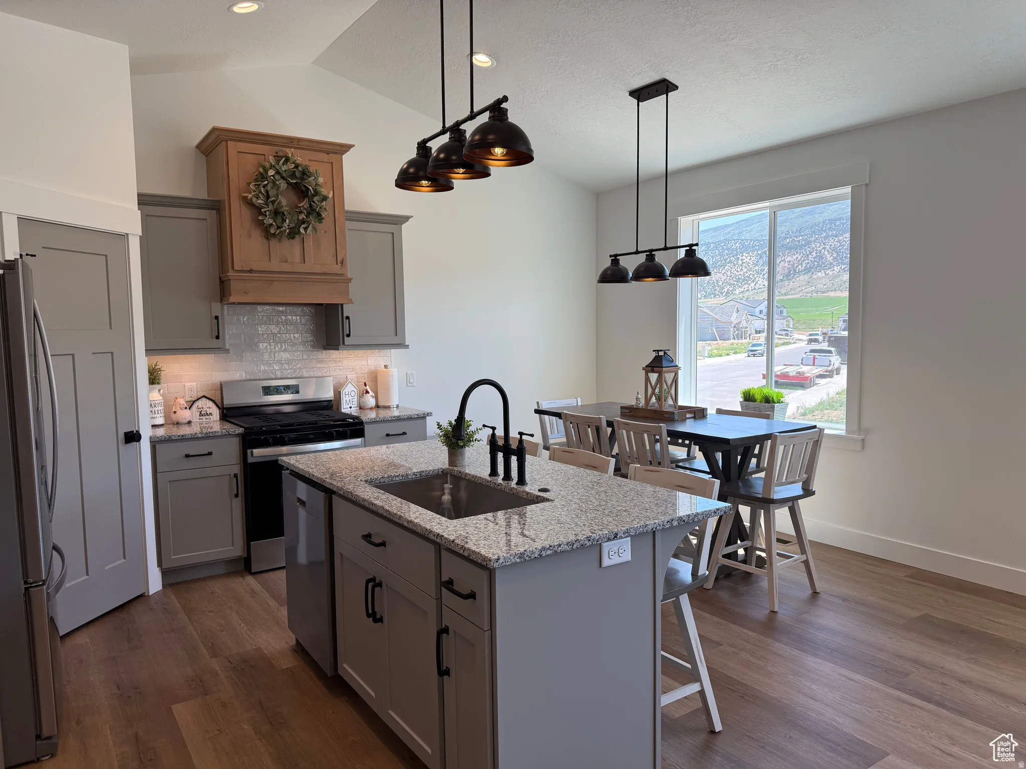 Kitchen with gray cabinets, light stone countertops, stainless steel appliances, a kitchen island with sink, and vaulted ceiling