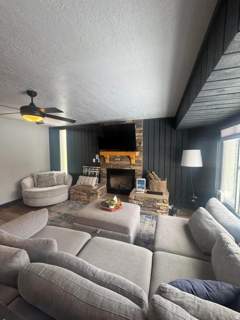 Living room featuring a stone fireplace, wood walls, a textured ceiling, ceiling fan, and wood finished floors