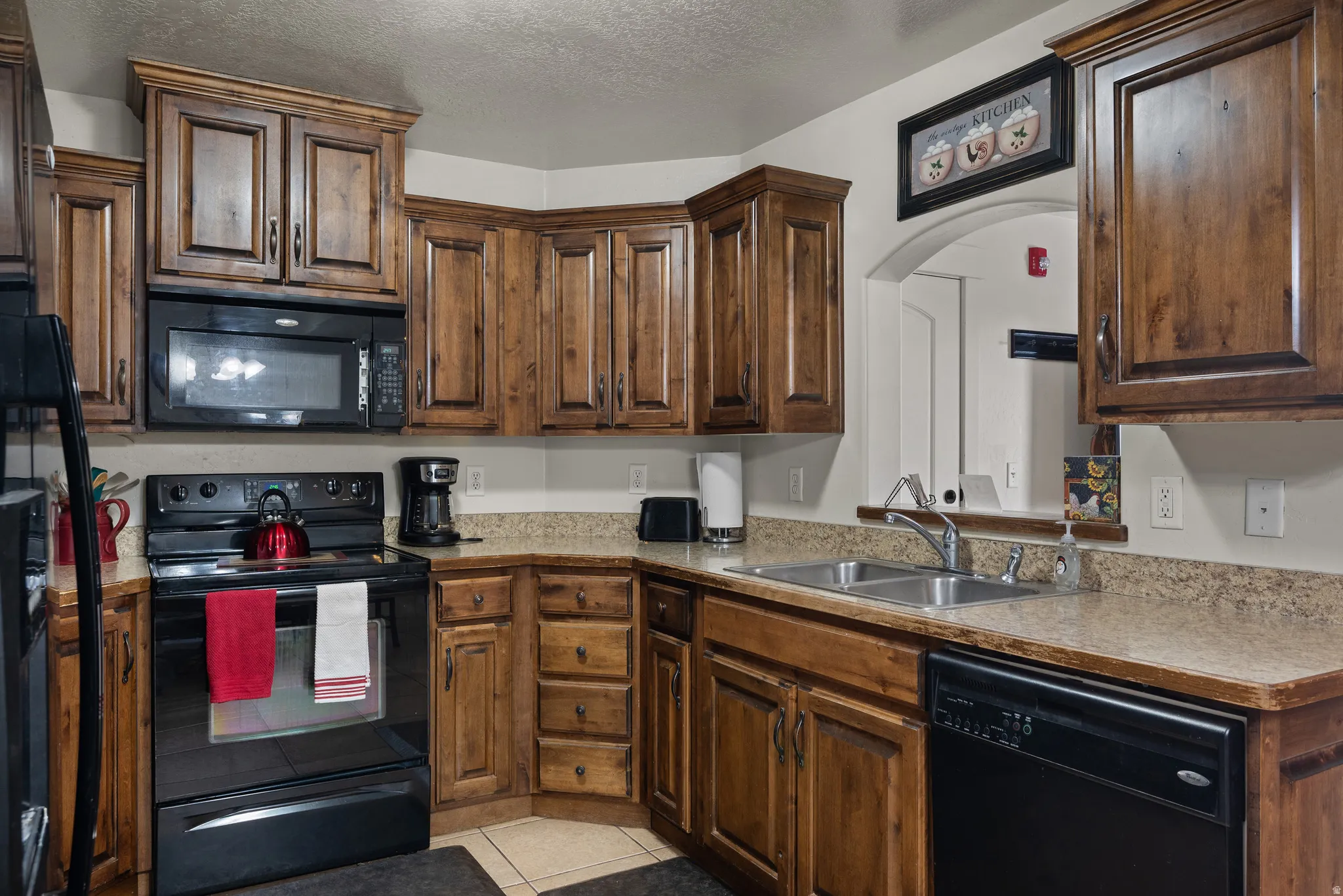 Kitchen featuring black appliances, a textured ceiling, light tile patterned flooring, light countertops, and dark wood finish cabinetry