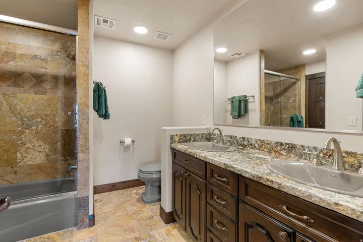 Bathroom with double vanity, bath / shower combo with glass door, and light stone finish flooring
