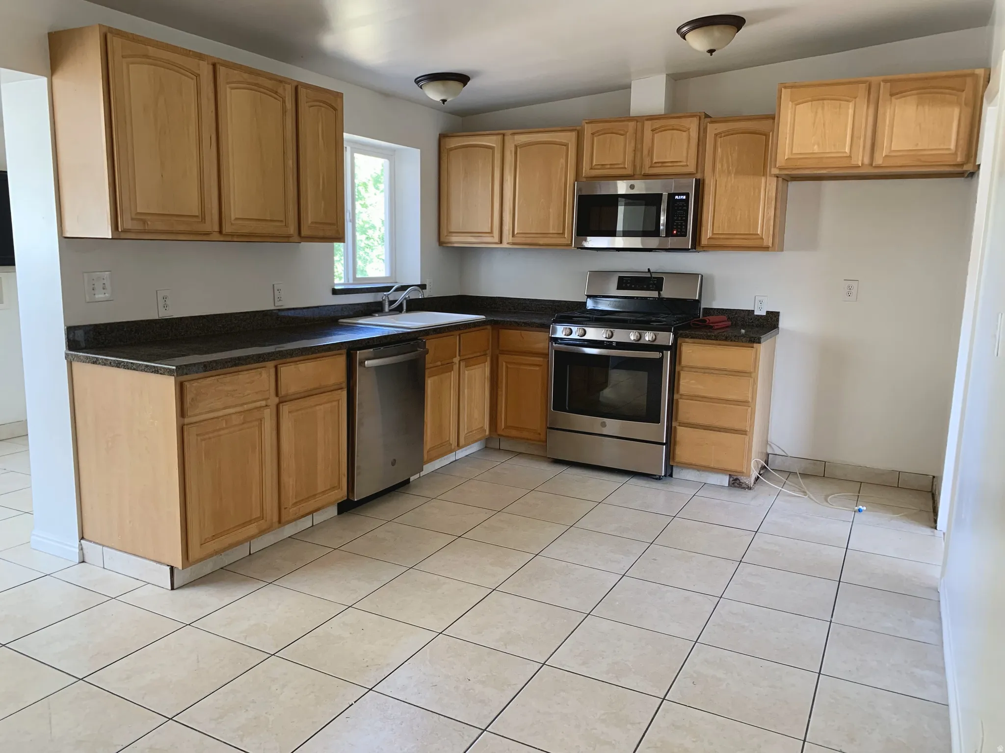Kitchen featuring stainless steel appliances, vaulted ceiling, light tile patterned flooring, wood finish cabinetry, and dark stone counters