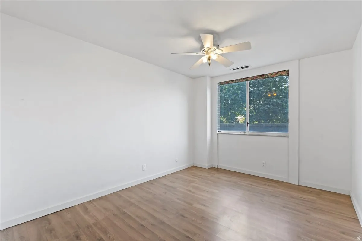 Spacious room featuring wood-finish flooring, a large window, a ceiling fan with integrated lighting, white baseboards, and painted walls