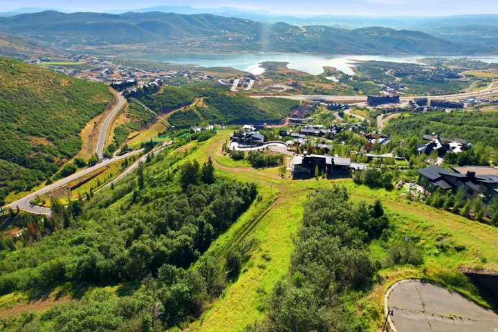 Expansive hillside setting with abundant greenery and distant water features, featuring a paved roadway and a cluster of residences