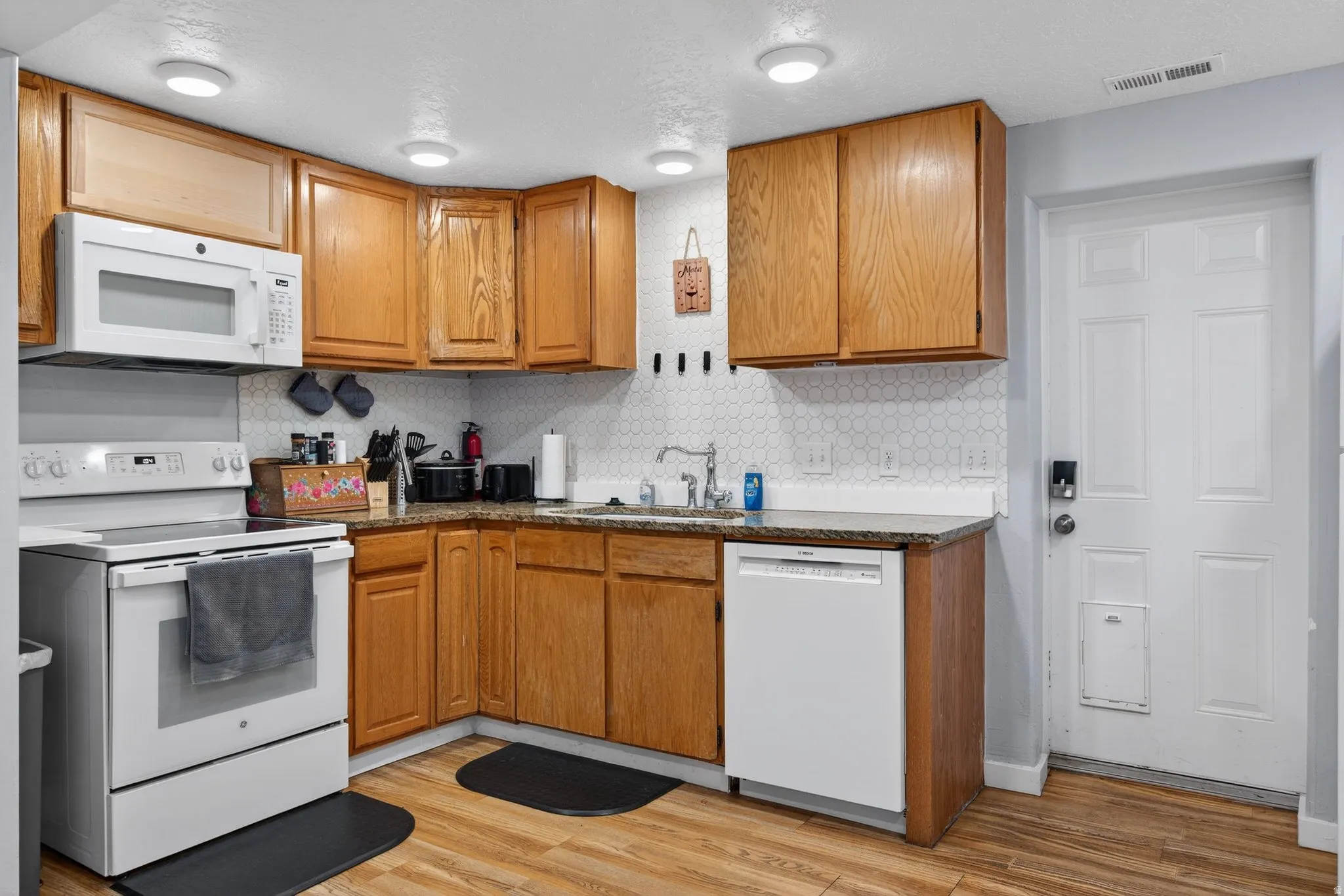 Kitchen with white appliances, wood finish cabinetry, dark stone countertops, light wood finished floors, and backsplash