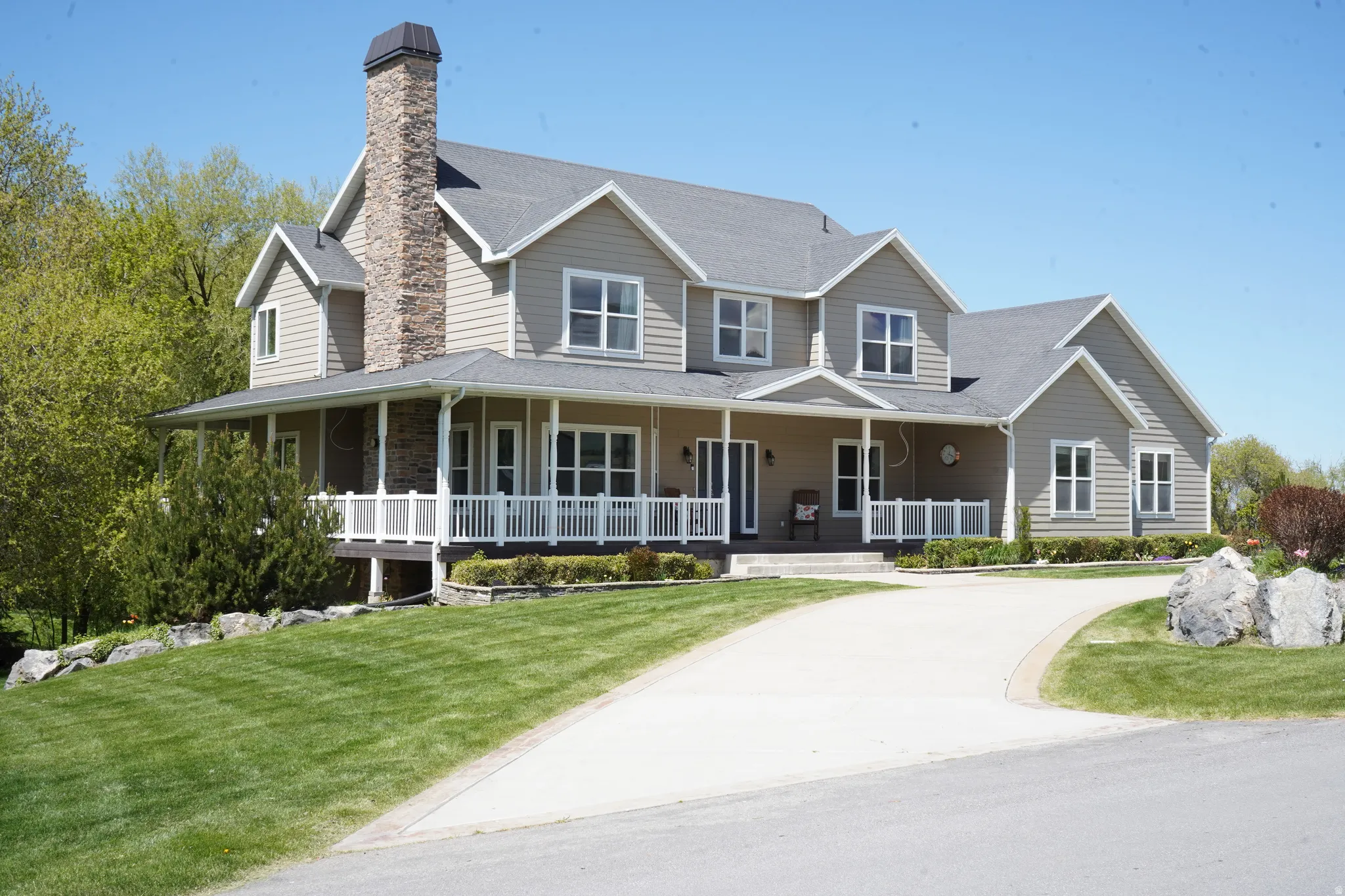 Expansive residence featuring a stone chimney, wrap-around covered porch with white railings, and tan horizontal siding