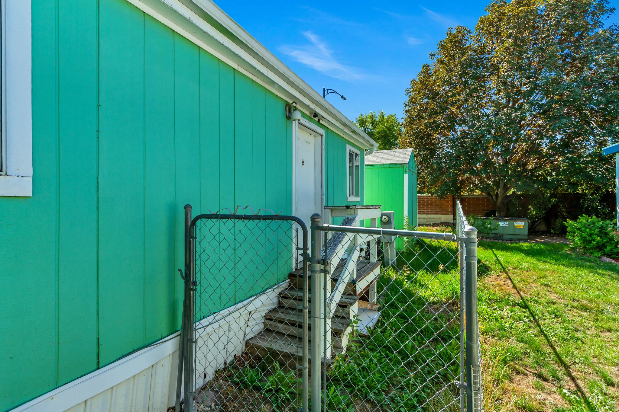 Exterior siding in a vibrant green finish, elevated wooden decking with stairs, a chain-link fence, and a separate storage shed