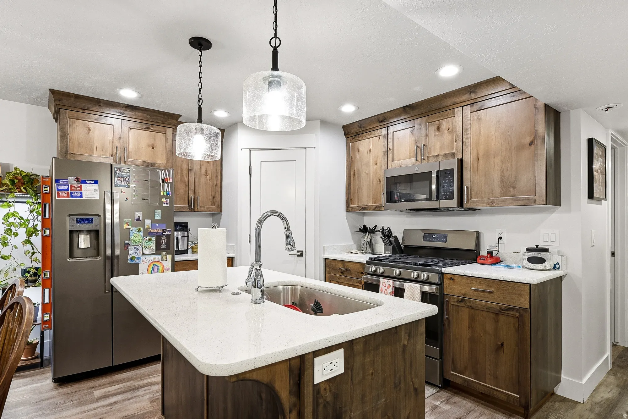 Kitchen featuring stainless steel appliances, light wood-style floors, a center island with sink, decorative light fixtures, and light stone countertops