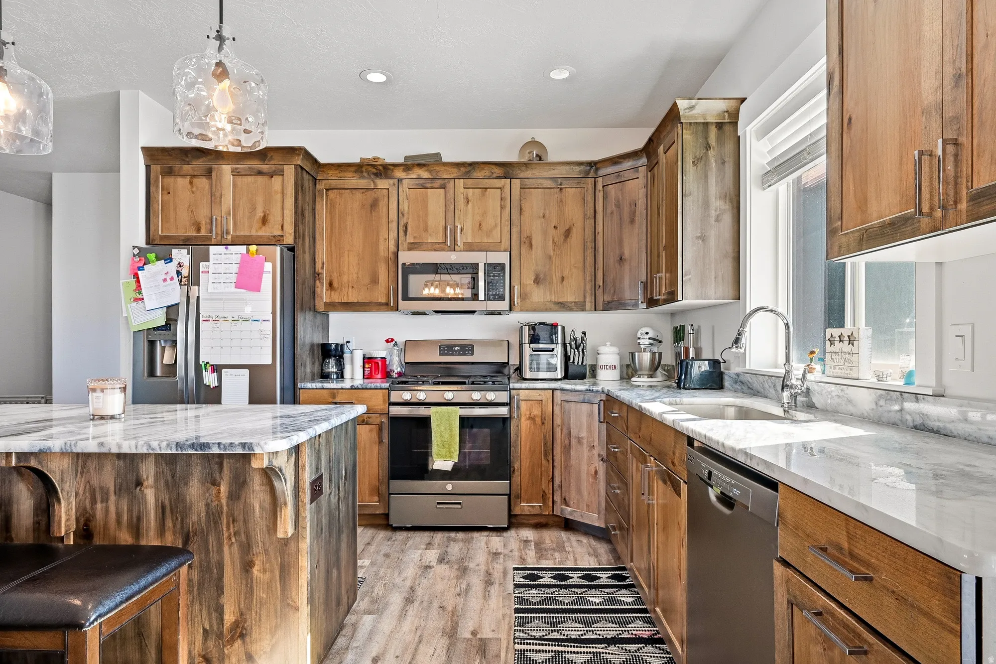 Kitchen featuring stainless steel appliances, light stone countertops, wood finish cabinetry, pendant lighting, and a kitchen bar
