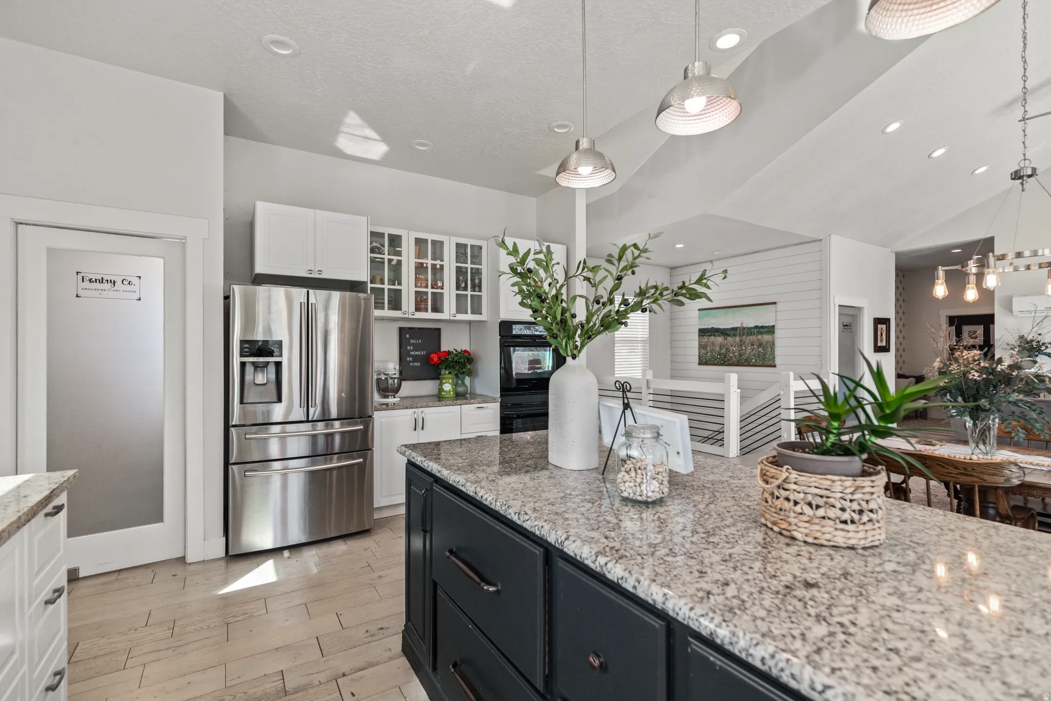 Kitchen featuring two tone cabinets, stainless steel fridge, glass insert cabinets, light stone counters, and decorative light fixtures