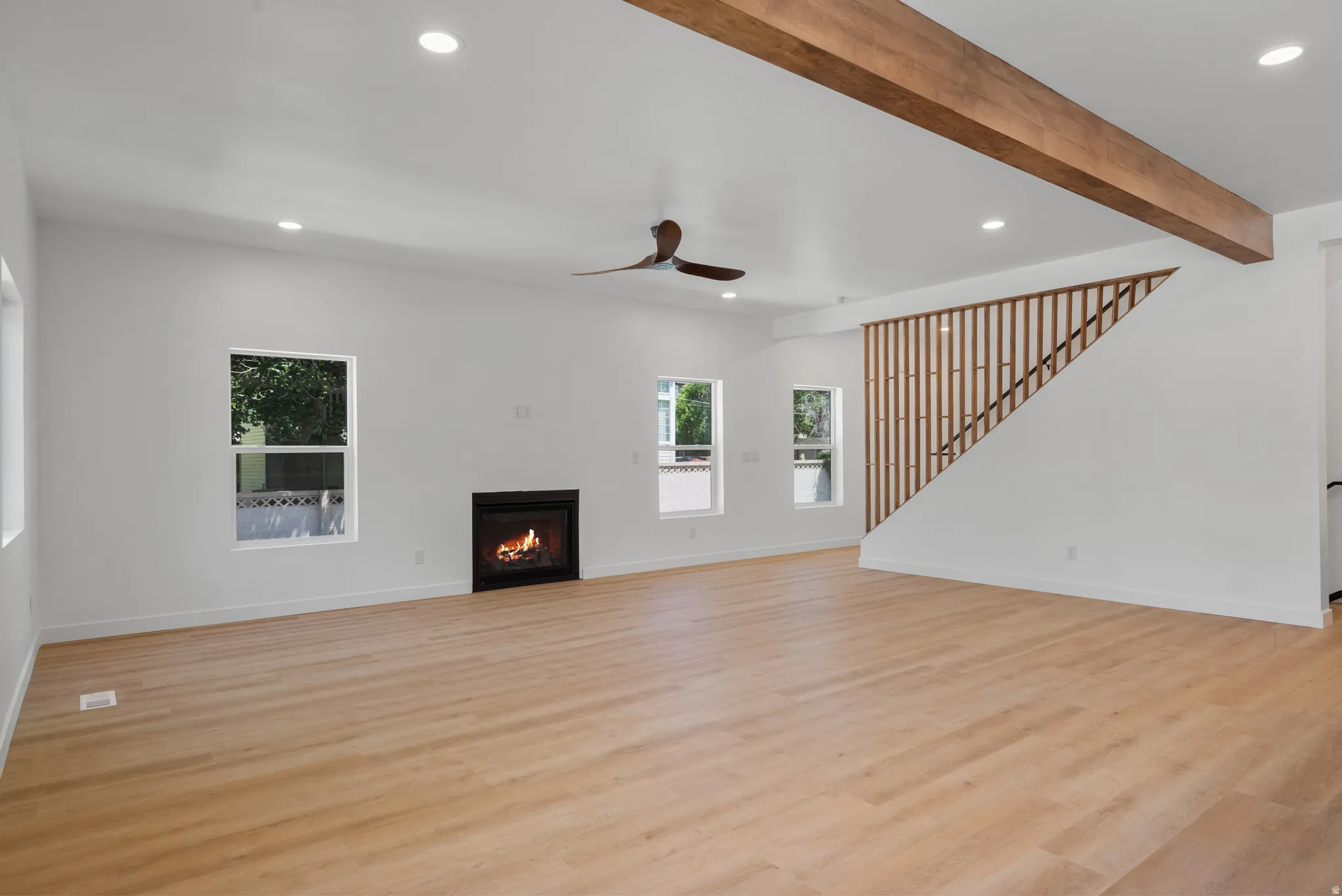 Spacious living area featuring wood-finish flooring, a modern fireplace, multiple windows, a decorative wood beam, and an open staircase with a slatted wood railing