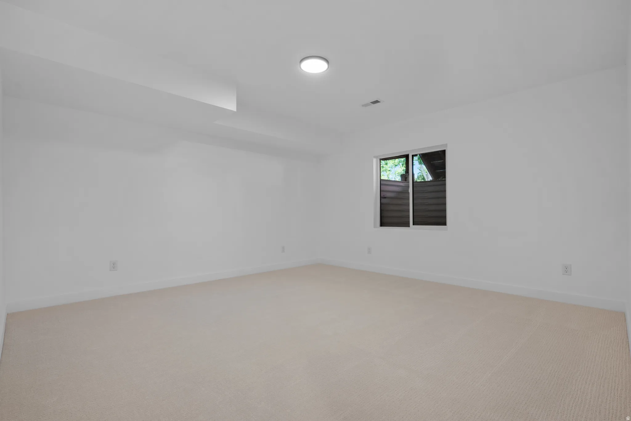 Spacious room featuring light-colored carpeting, white painted walls, and a single window with dark framing