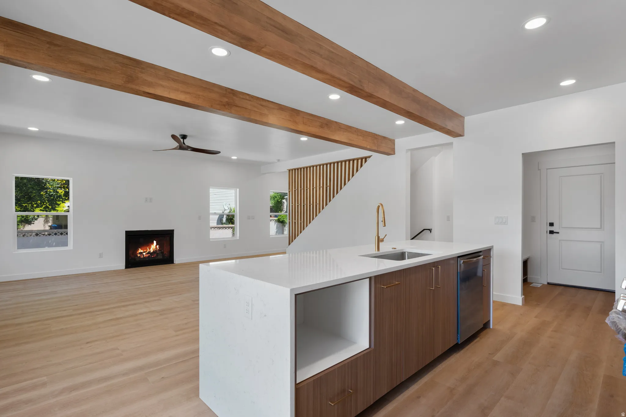 Open-concept living area featuring a kitchen island with a gold-finish faucet and integrated sink, exposed wood beams, wood-finish flooring, a contemporary fireplace, and recessed lighting