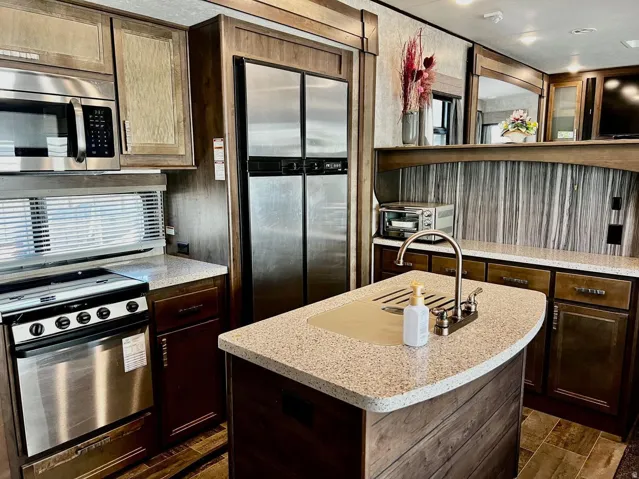 Kitchen with stainless steel appliances, a center island with sink, light stone counters, wood tiled floors, and dark wood finish cabinetry