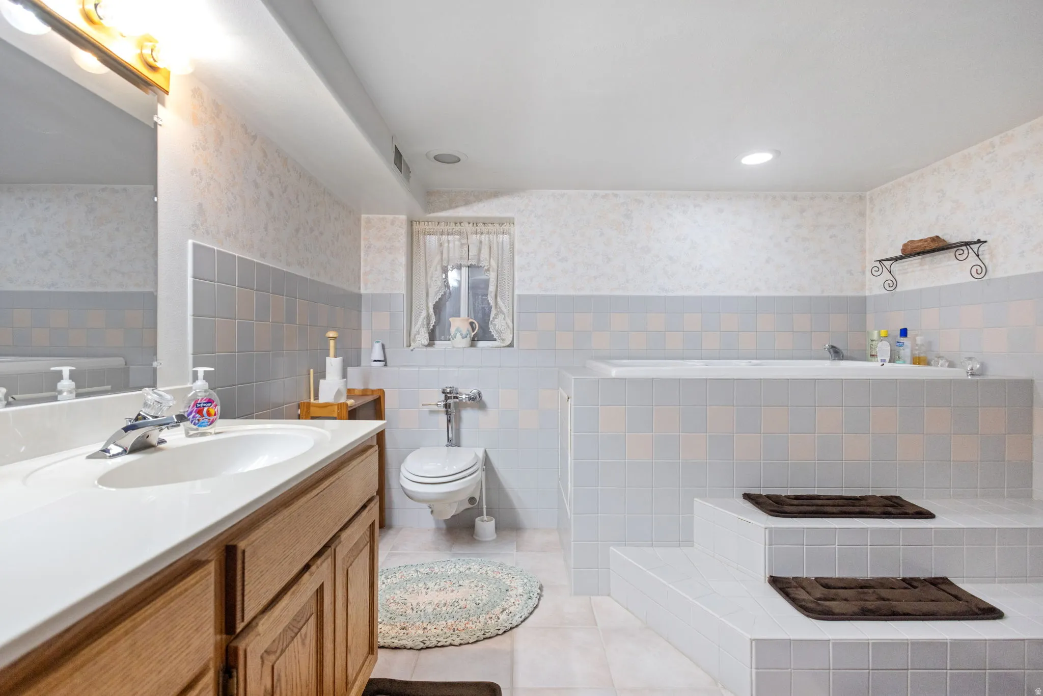 Full bathroom with wallpapered walls, a wainscoted wall, tile walls, light tile patterned floors, and vanity