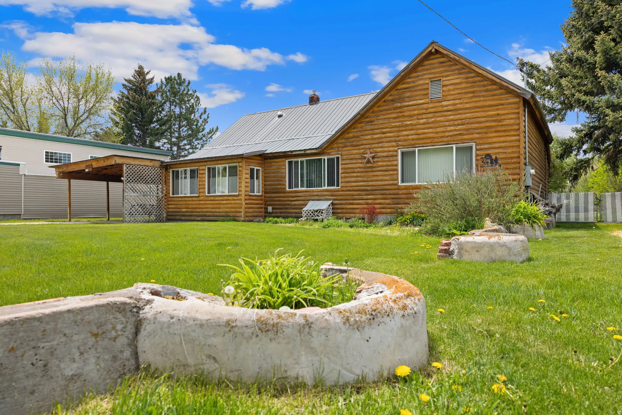 Log cabin style exterior featuring a metal roof, multiple windows, an attached carport with a lattice side, and a spacious lawn with mature trees