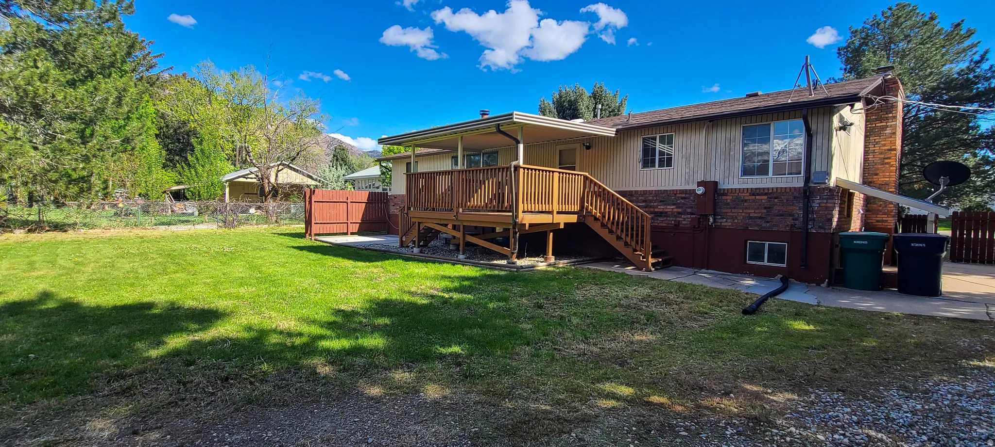 Rear exterior featuring a large covered wooden deck with railing and stairs,  brick foundation accents, and a spacious grassy yard