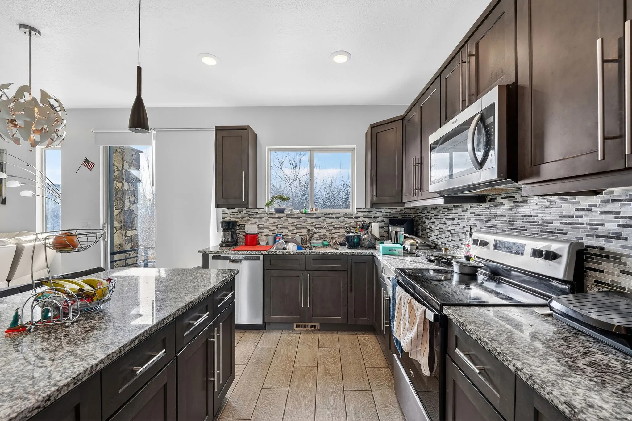 Kitchen with stainless steel appliances, dark wood finish cabinets, light stone countertops, light wood-style floors, and pendant lighting