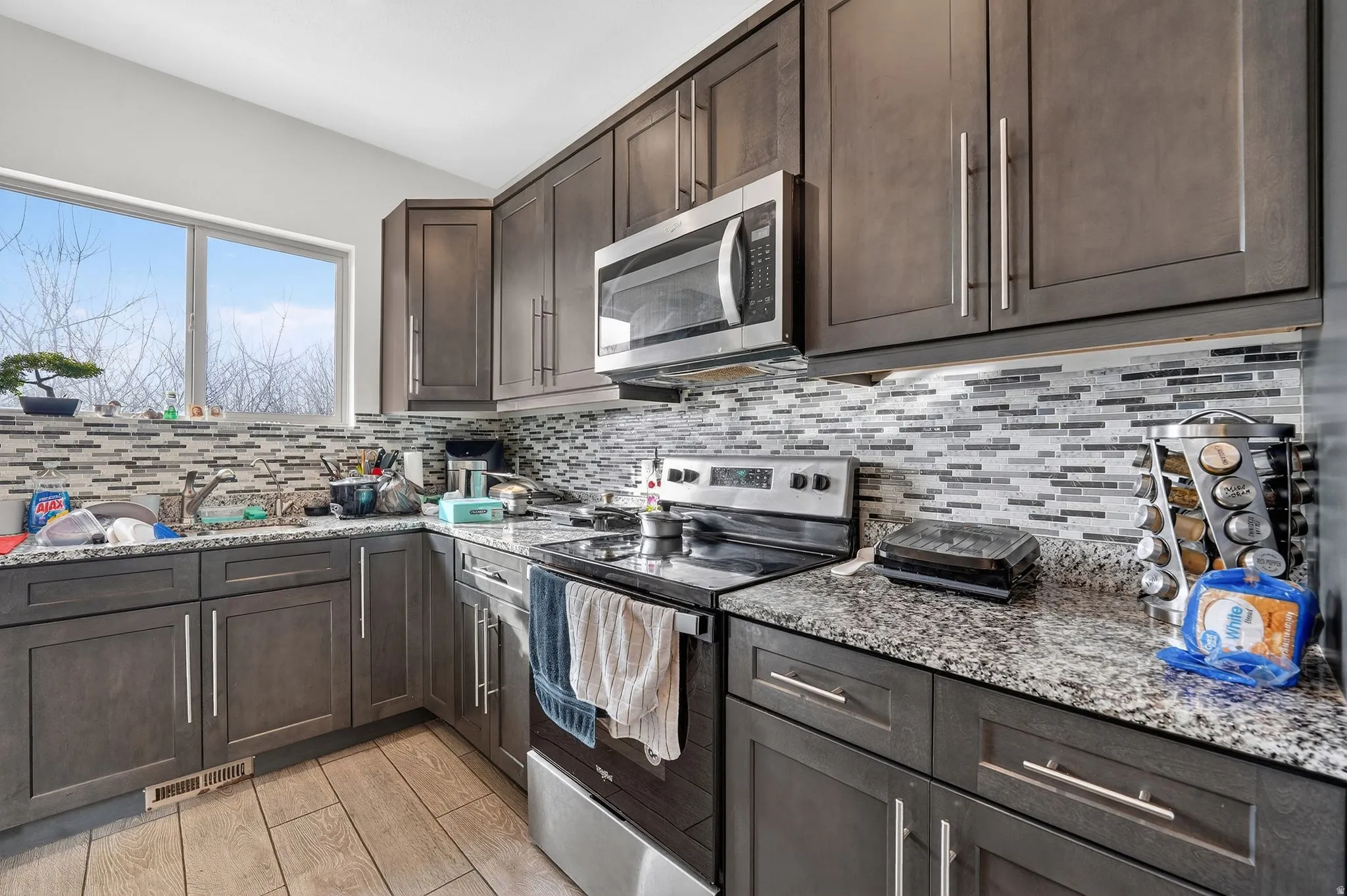Kitchen with stainless steel appliances, dark wood finish cabinets, light stone countertops, light wood-style flooring, and backsplash
