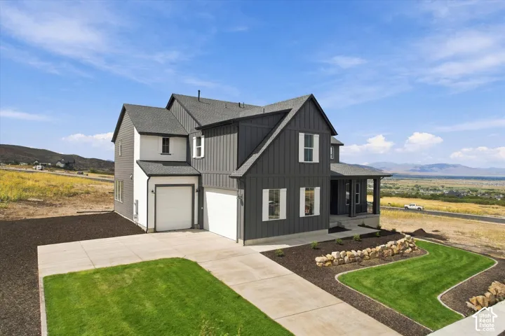 Two-story farmhouse-style exterior featuring vertical board-and-batten siding, contrasting white and charcoal finishes, dual-car garage, stacked stone retaining wall, and mountain backdrop