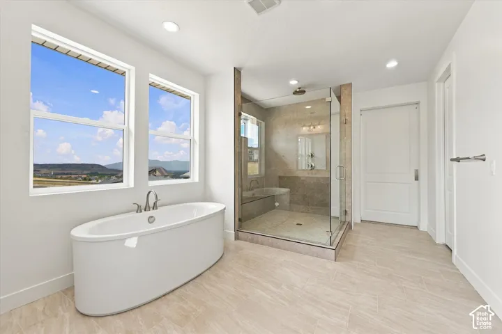 Freestanding oval bathtub with deck-mounted faucet, walk-in glass-enclosed shower featuring tiled surround and bench seating, tile flooring, and large windows with mountain scenery