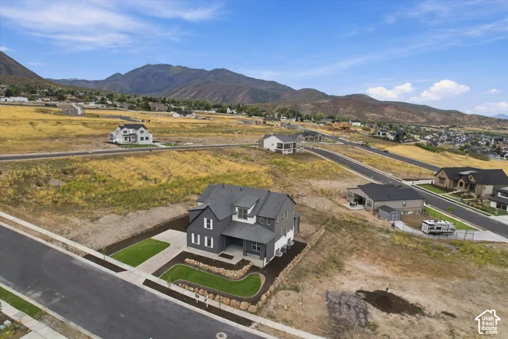 Aerial perspective featuring a multi-story residence with gray siding, dark shingled roof, mountain backdrop, and landscaped front yard with stone retaining wall