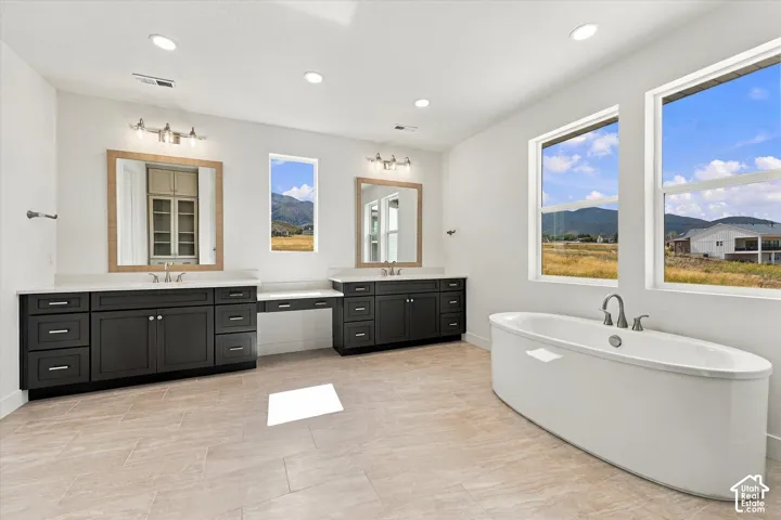 Double vanity featuring dark cabinetry, solid surface countertops, inset sinks, and dual framed mirrors, paired with a freestanding soaking tub, ceramic tile flooring, and mountain backdrop