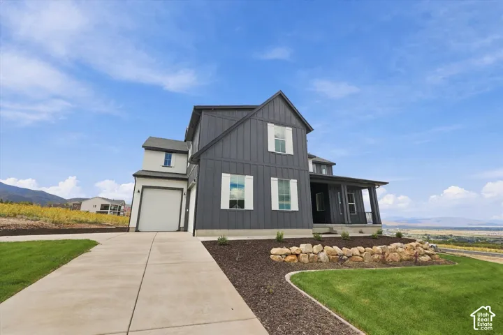 Two-story exterior featuring board-and-batten siding, contrasting window shutters, a covered porch, a concrete driveway, and natural stone landscaping elements