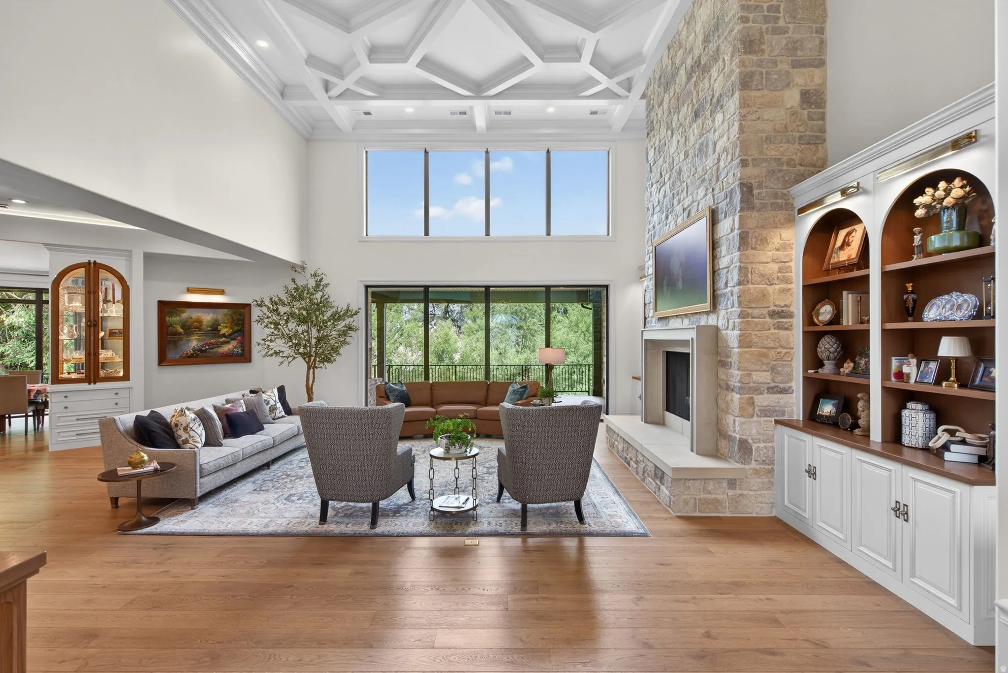 Spacious living area featuring 19 ft. ceiling with intricate geometric detailing, a stone-clad fireplace, built-in shelving with arched accents, wood-finish flooring, and large windows providing abundant natural light