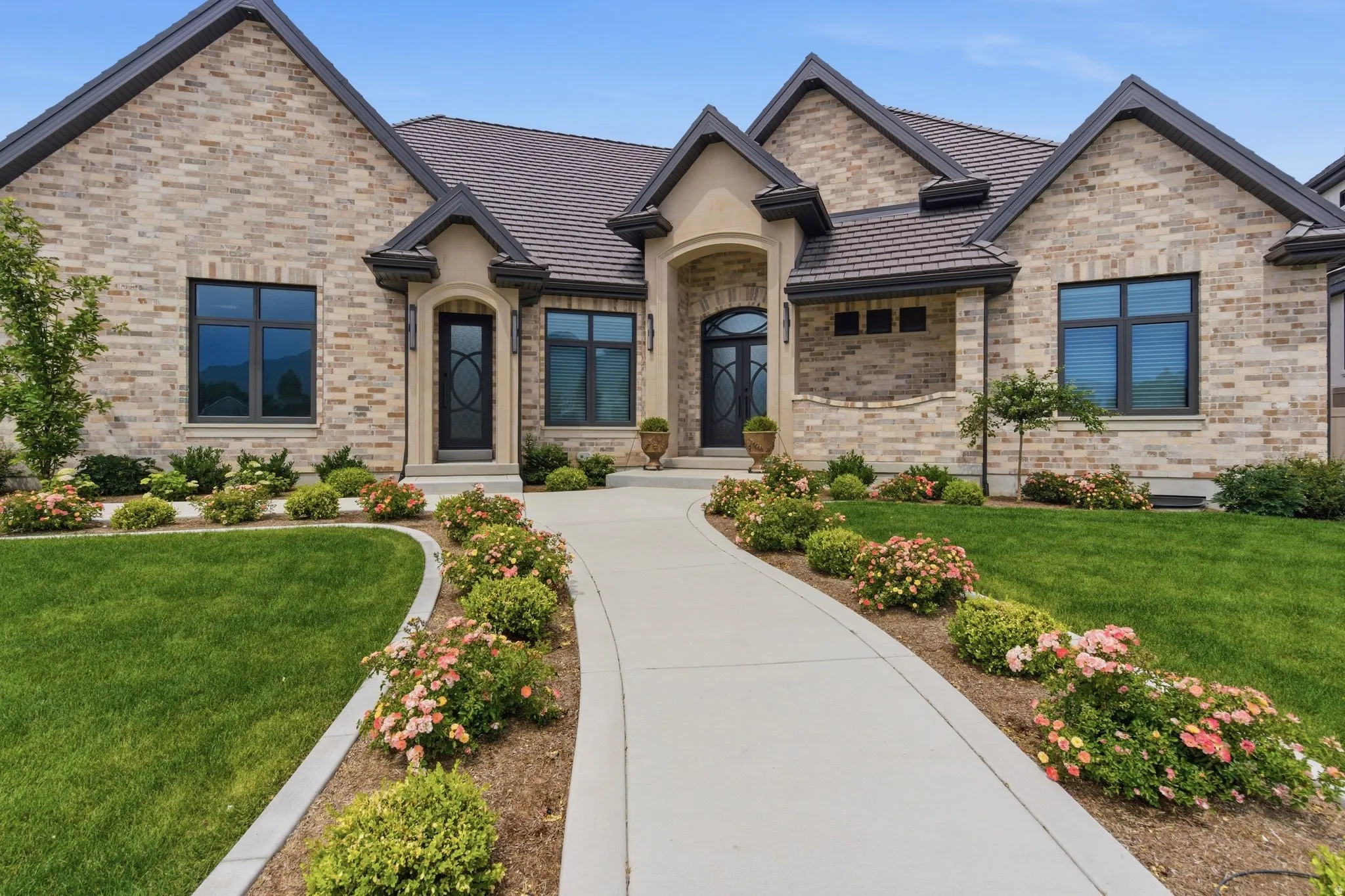 Inviting exterior featuring a curved concrete walkway, brick facade, arched limestone entryways, dark-framed windows, and beautifully landscaped with shrubs and flowering plants