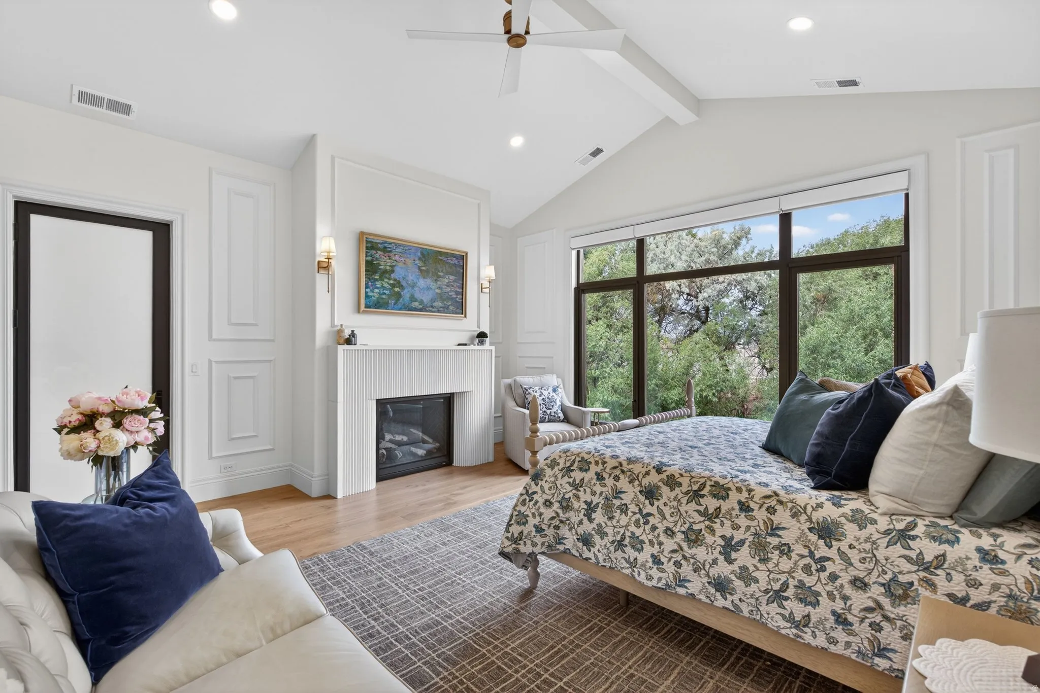Primary Bedroom Bright interior featuring vaulted ceilings with beams,  ceiling fan, large window with dark trim, a built-in fireplace with fluted detailing, and wood-finish flooring
