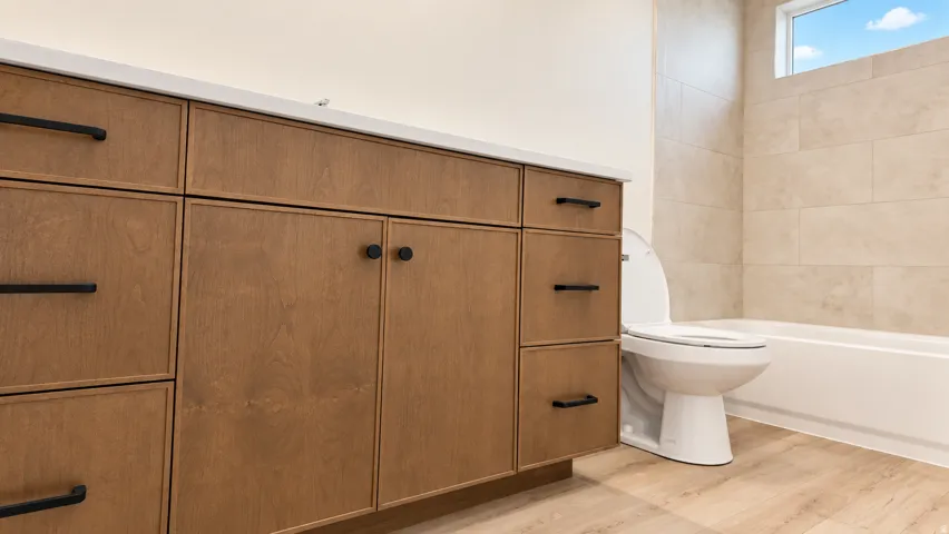Wood-finish vanity with matte black hardware, white countertop, tile shower surround, and a soaking tub