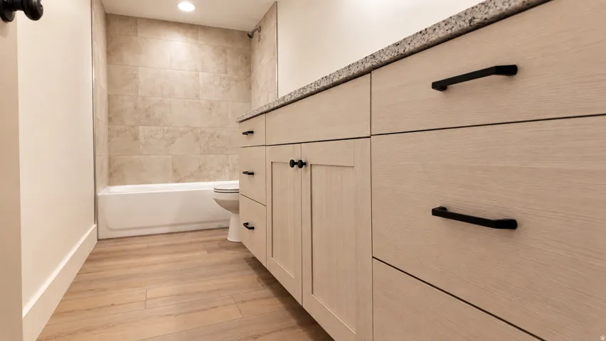 Light wood-finish flooring, built-in vanity with light wood-finish cabinetry, black hardware, speckled countertop, and a tiled shower-tub combination