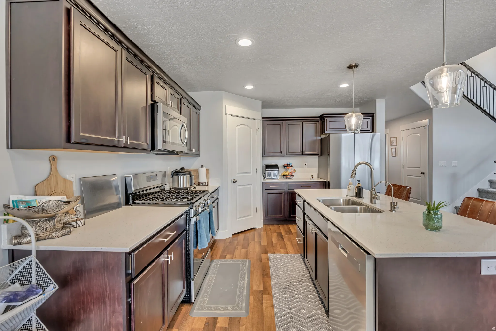 Kitchen featuring stainless steel appliances, dark wood finish cabinetry, a kitchen island with sink, decorative light fixtures, and light wood finished floors