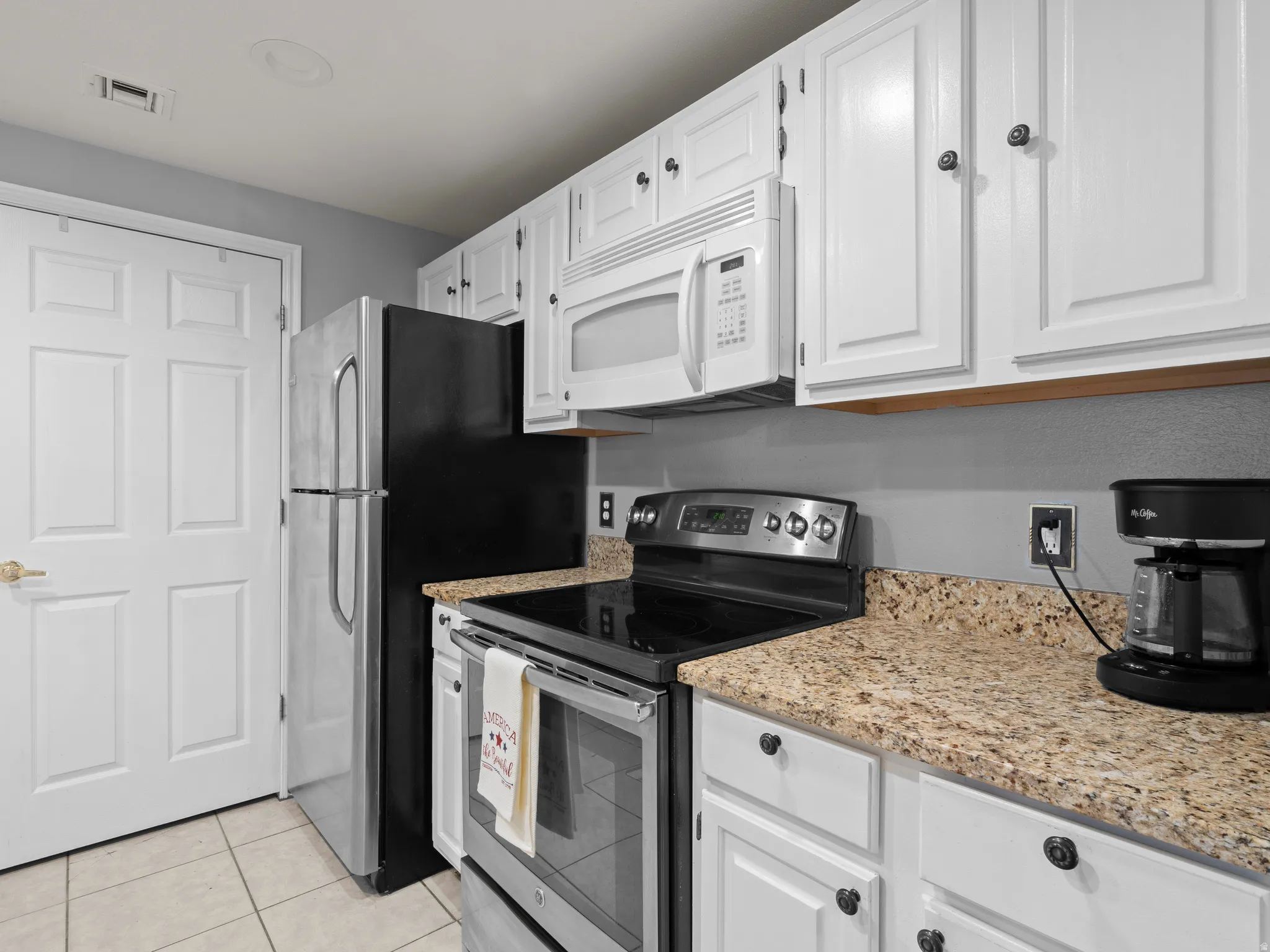 Kitchen featuring stainless steel electric range oven, white microwave, white cabinets, light stone countertops, and light tile patterned floors