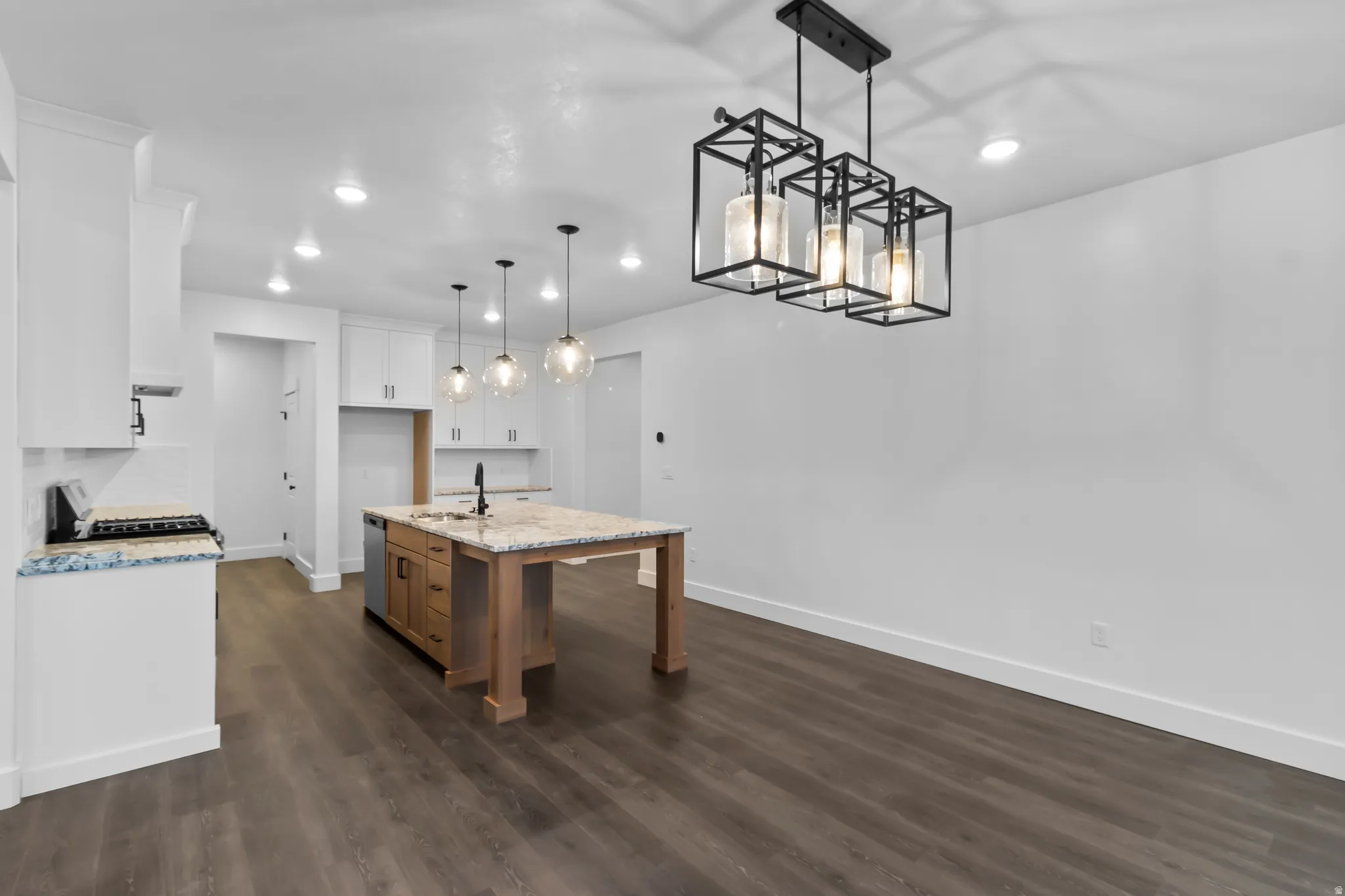 Two tone kitchen with two tone cabinetry, a center island with sink, pendant lighting, light stone counters, and dark wood-type flooring