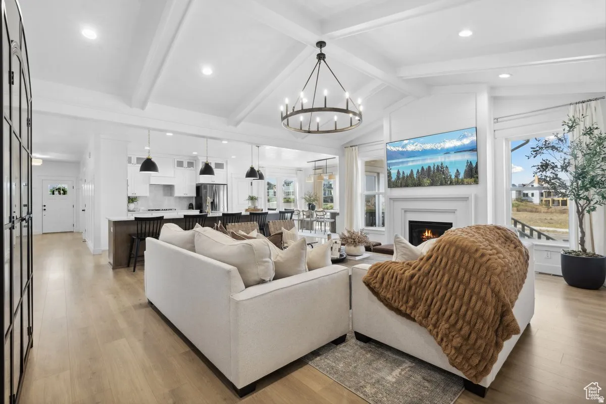 Living room featuring a warm lit fireplace, light wood finished floors, a chandelier, healthy amount of natural light, and beamed ceiling
