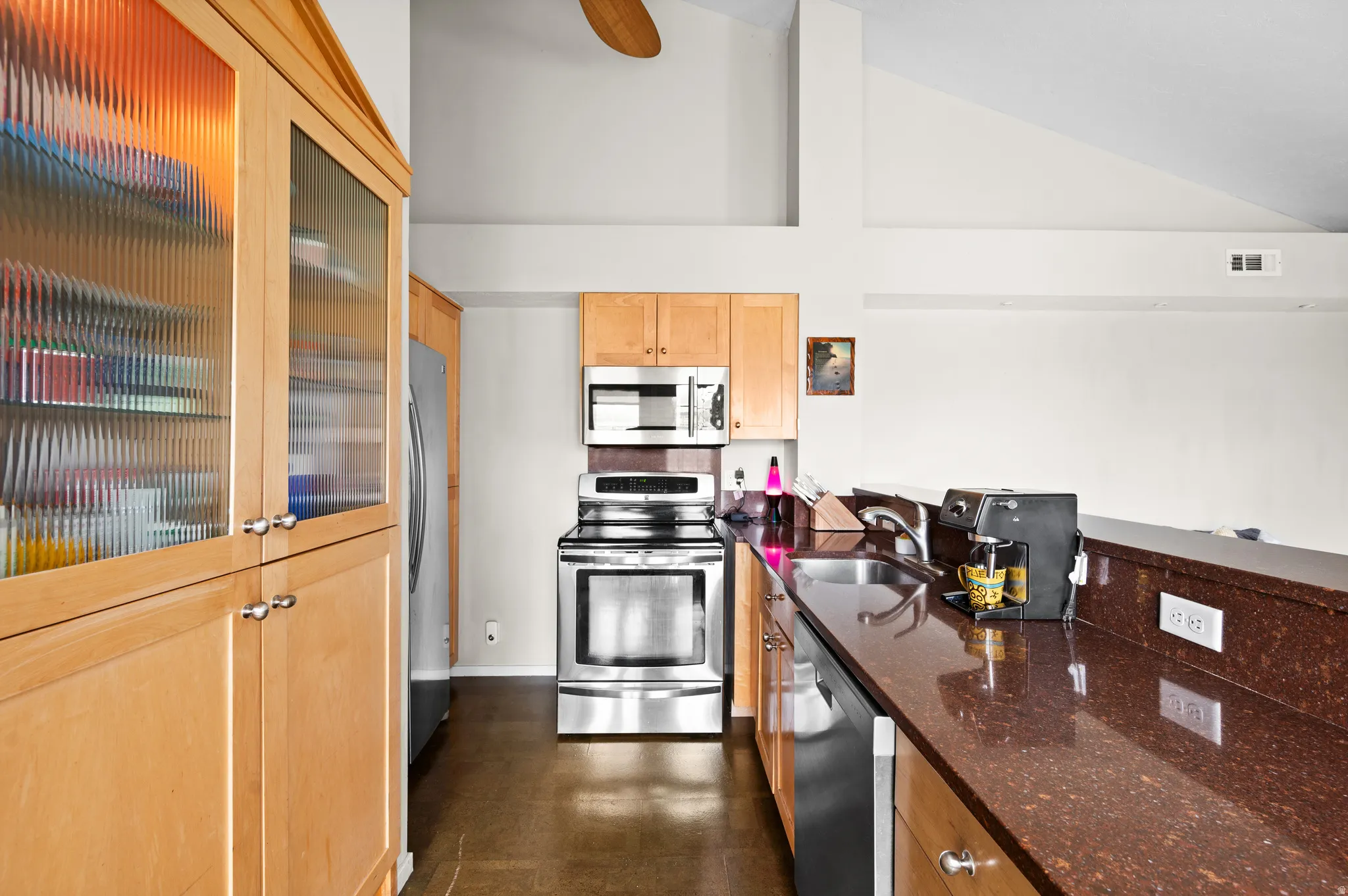 Kitchen with glass insert cabinets, stainless steel appliances, dark stone countertops, light wood finish cabinets, and vaulted ceiling