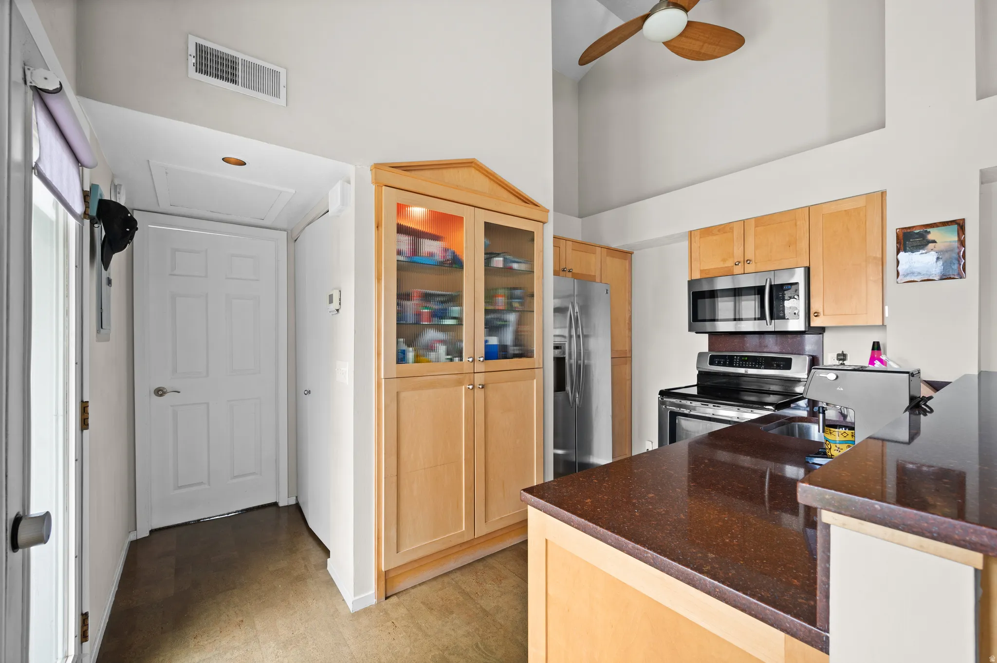 Kitchen with light wood finish cabinets, stainless steel appliances, glass fronted cabinets, dark stone counters, and ceiling fan