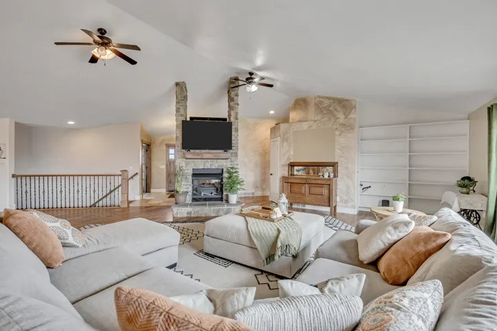 Living area with wood finished floors, lofted ceiling, a stone fireplace, ceiling fan, and built in shelves
