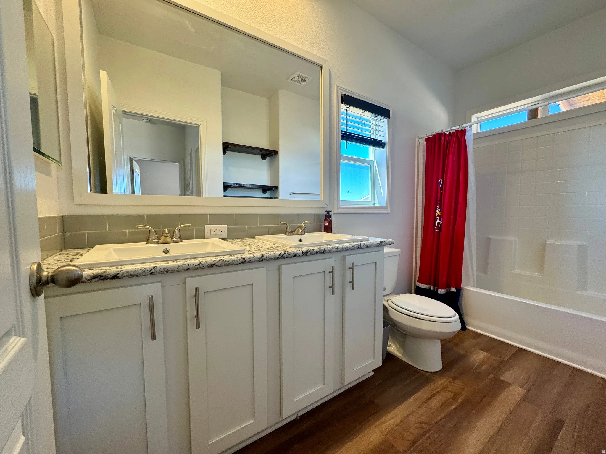 Full bathroom with double vanity, dark wood-style flooring, tasteful backsplash, and shower / tub combo with curtain