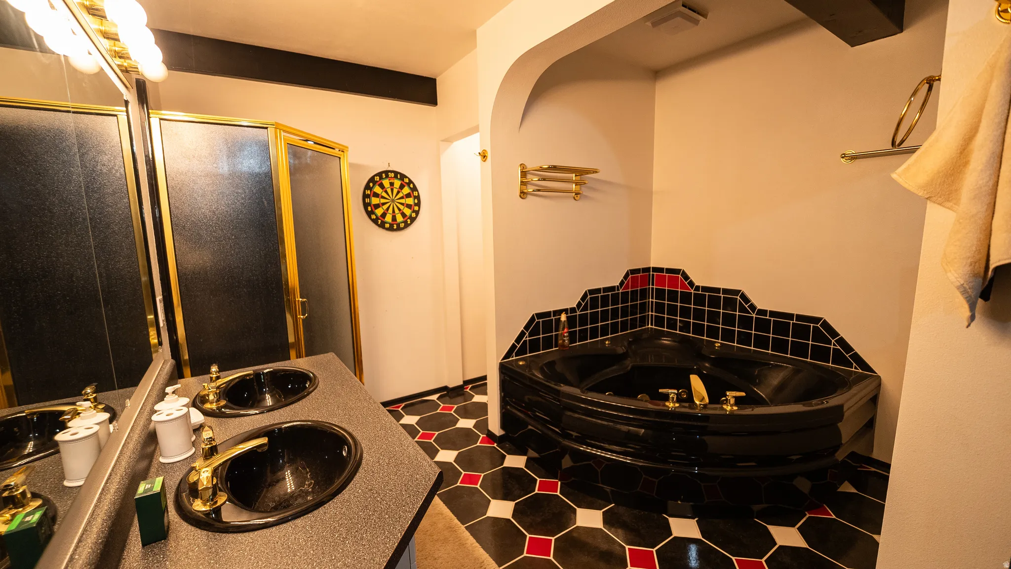 Bathroom featuring a corner soaking tub with black and red tiled surround, black dual sinks with brass fixtures, and a glass-enclosed shower with brass framing