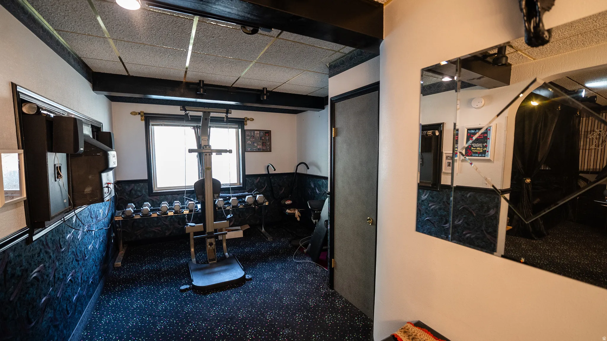 Recreational room featuring textured wall panels, a window with horizontal blinds, and a speckled carpet flooring