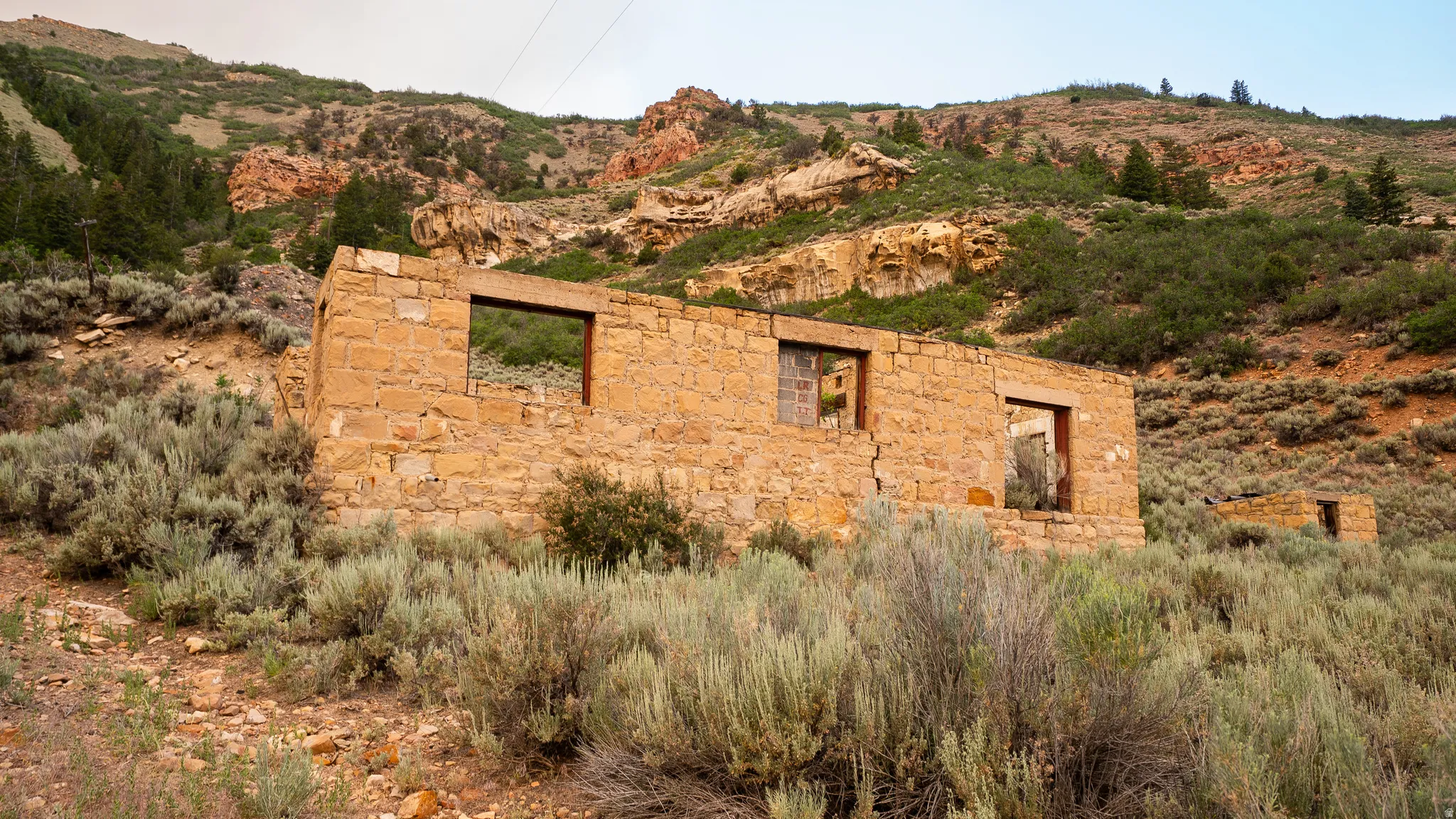 Stone foundation and wall remnants set into a natural, sloped terrain with abundant sagebrush and distant rock formations