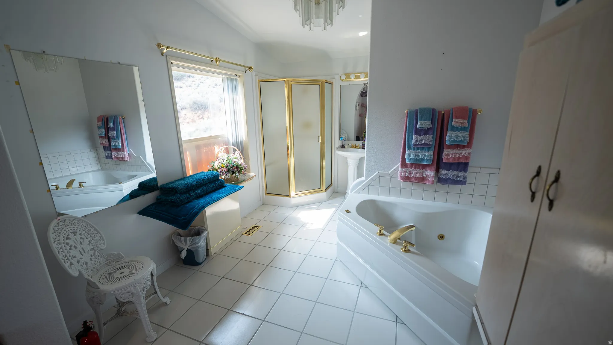 Spacious bathroom featuring a corner jetted tub, a separate corner shower with gold-toned framing, a pedestal sink, and white square floor tiling