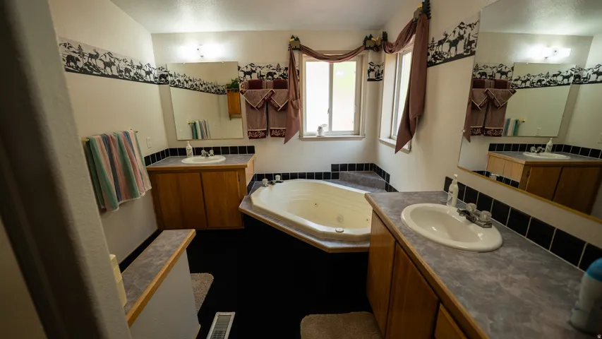 Bathroom featuring a corner jetted tub with tiled surround, dual vanity sinks with wood cabinetry and laminate countertops, and wall-mounted mirrors