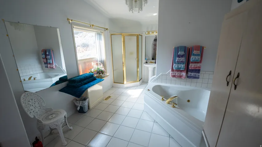 Spacious bathroom featuring a corner jetted tub, a separate corner shower with gold-toned framing, a pedestal sink, and white square floor tiling