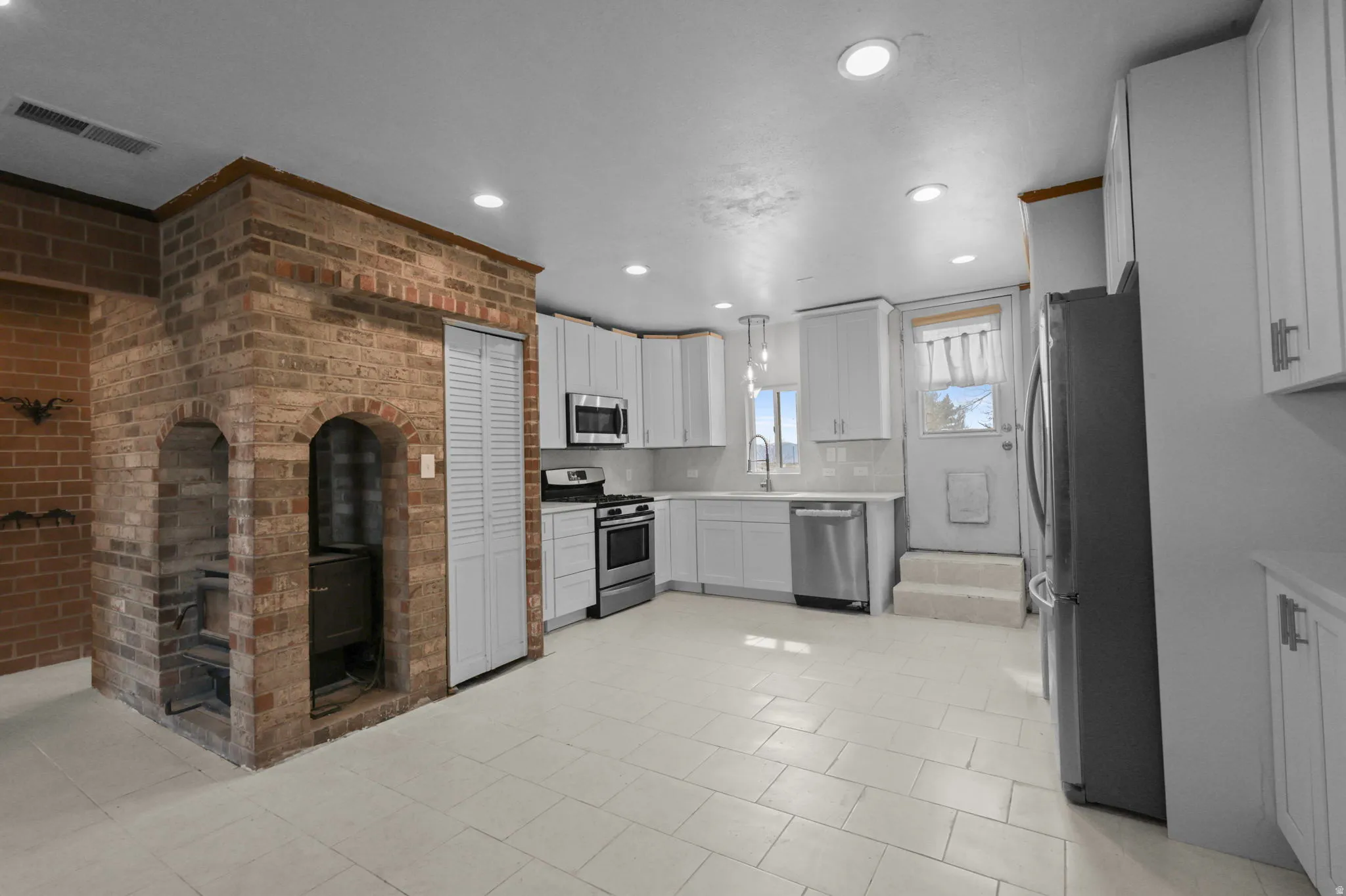 Kitchen with stainless steel appliances, white cabinets, brick wall, hanging light fixtures, and tasteful backsplash