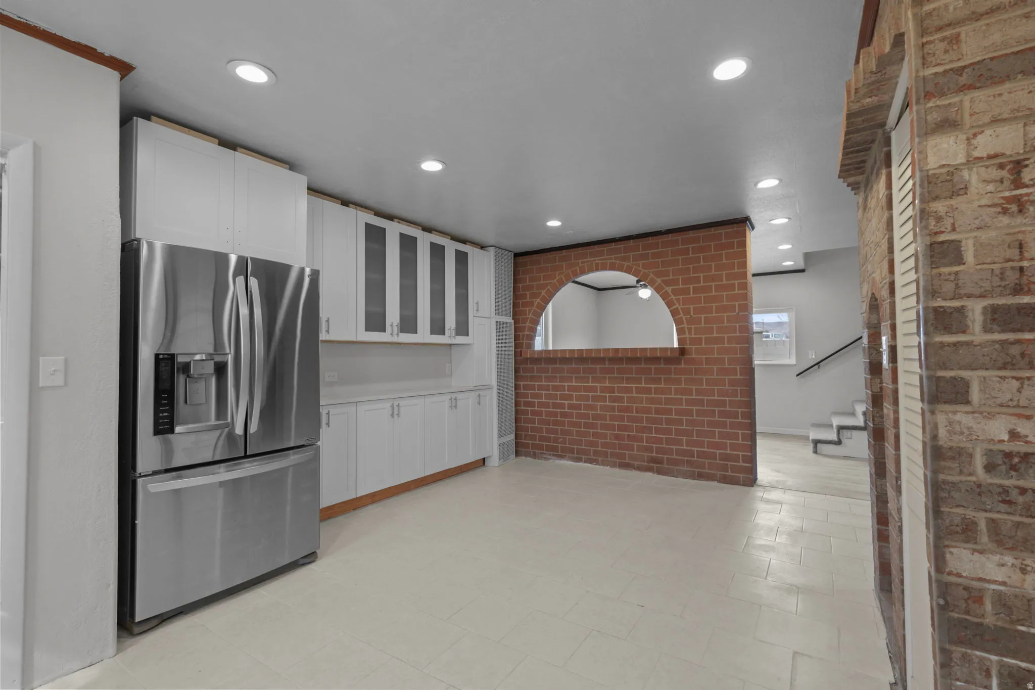Kitchen featuring brick wall, stainless steel fridge, glass fronted cabinets, white cabinetry, and light countertops