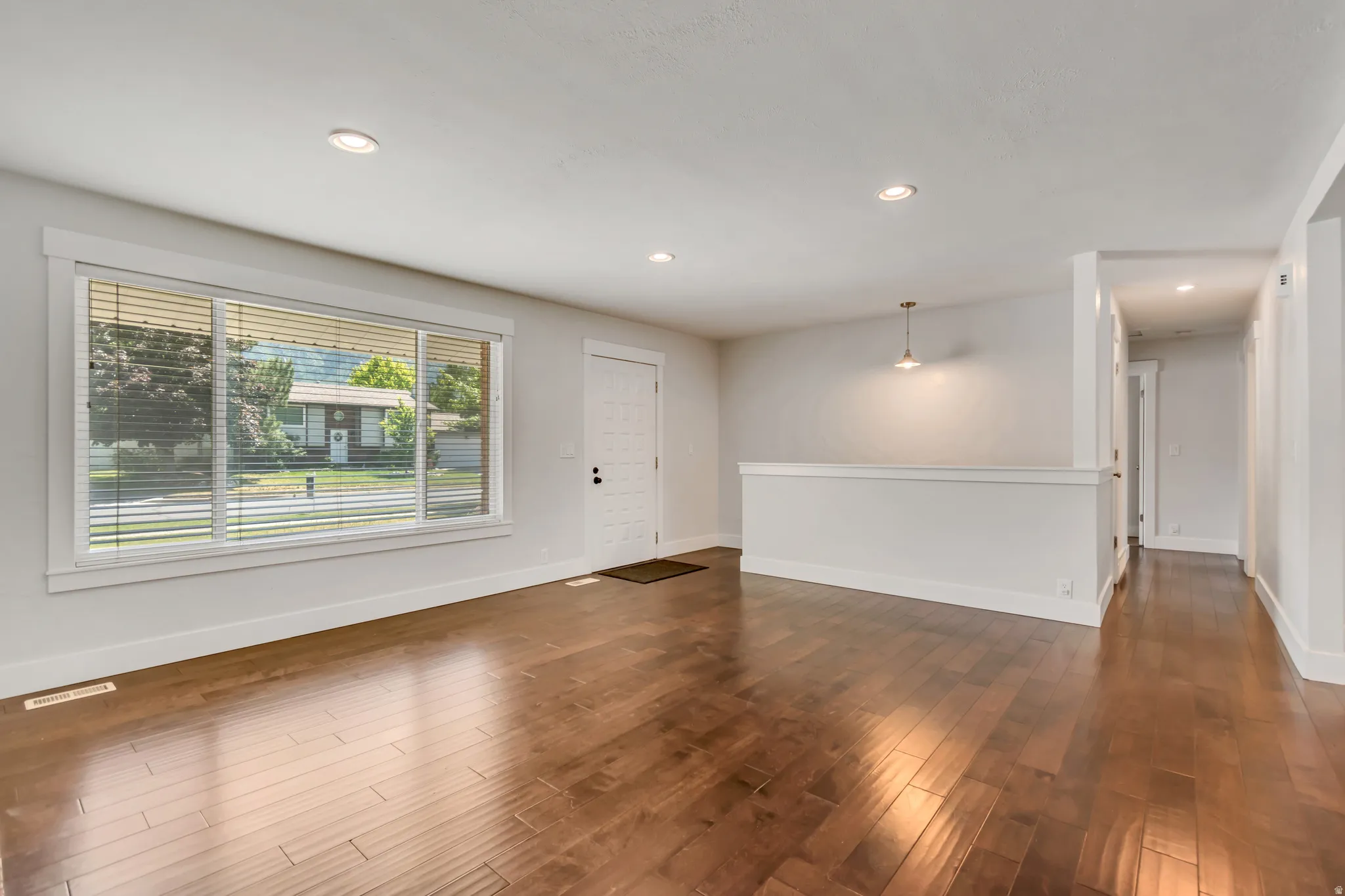 Spacious living area featuring wood-finish flooring, recessed lighting, and a prominent front window with horizontal blinds