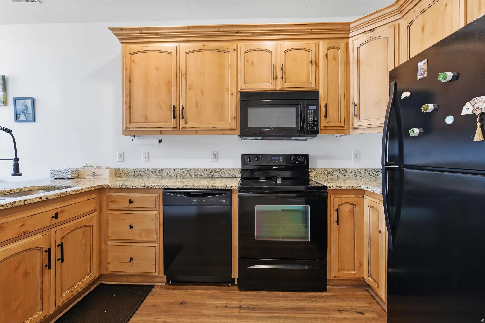 Kitchen with black appliances, light stone countertops, light wood-type flooring, and light wood finish cabinetry